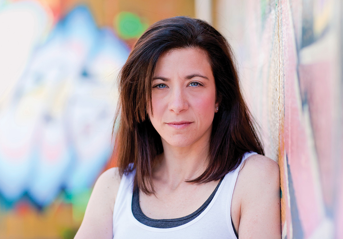 A brown-haired woman staring into the camera in front of graffitied walls