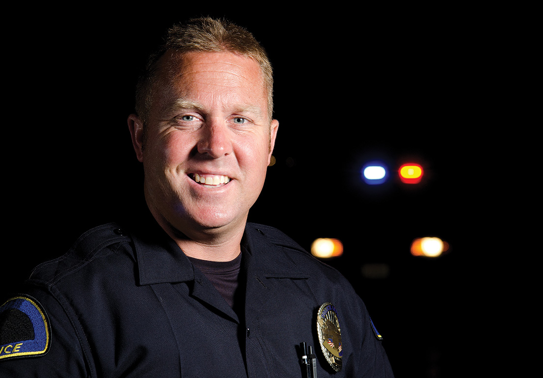 A police officer smiling, police car lights shine behind him